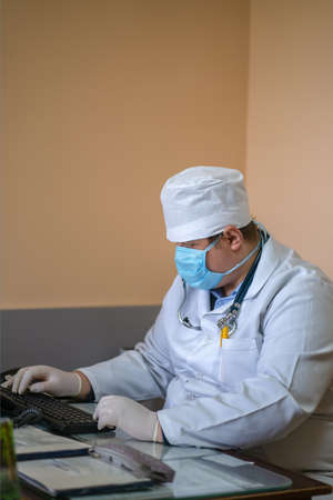 Masked GP doctor using computer. General practitioner works with computer application sitting at desk in his office. City Hospital. May, 2020, Brovary, Ukraine.のeditorial素材