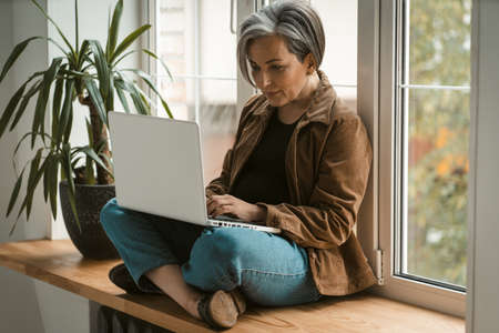 Serious mature woman in casual uses laptop computer sitting crossed legs on wooden window sill....