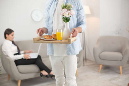 Man bringing his wife food. Cropped picture of man holding food tray. Cropped shot of male wearing white clothes holding up tray with breakfast food on it. Woman sits at sofa on blurred background.の写真素材