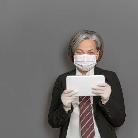 Caucasian businesswoman in protective mask works digital gadget. Gray haired lady looks at camera...
