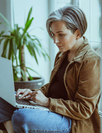 Elegant Mature woman works laptop sitting on sill of window. Side view of modern silver haired Lady in casual typing on computer keyboard.の写真素材