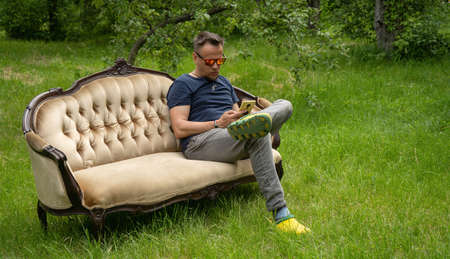 Middle-aged man uses mobile phone resting on light sofa in garden. Sofa is on green grass outdoors....