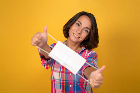 Smiling woman holds protective mask in front of her. Cut out on yellow background. Quarantine...