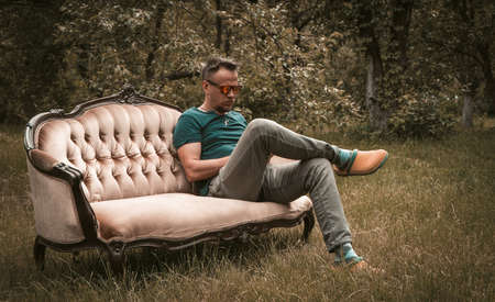 Resting man in grey jeans sitting on a sofa in the middle of garden. High quality photo