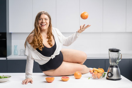 Girl with obese problem. Young sexy chubby white girl in black swimsuit, white shirt at modern kitchen table. juggling fruits in hand. Trying to loose weight fast. Fat barefoot girl low carb diet.の写真素材