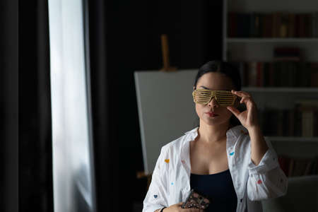 Asian middle aged woman is disco shades or party glasses with easel behind her. Mature serious...