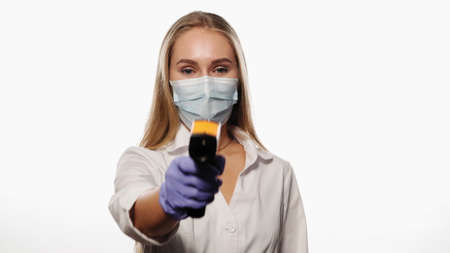 Doctor or nurse pointing sensor thermometer on camera wearing protective mask white medical hospital robe. Young blond haired nurse checking on patients or coworker temperature. 4k UHD.の写真素材