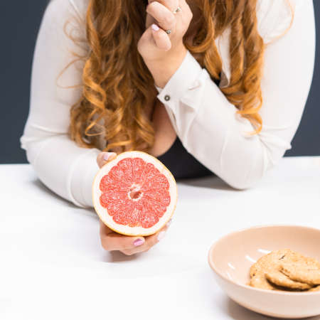 Fat young woman holding a fresh grapefruit in hand standing leaned on a kitchen table at home with...