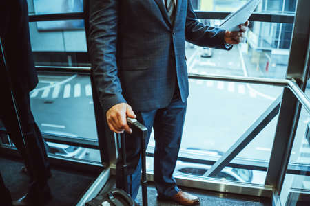 No face visible. Businessman looking at documents wearing a suit standing next to the window with suitcase next to him in the modern office with street view. Man holding a papers next to the window.の写真素材