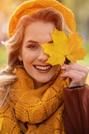Happy young girl cover her eye with a yellowed leaves in yellow knitted beret with autumn leaves in hand and fall yellow garden or park. Beautiful smiling young woman in autumn foliage.の写真素材