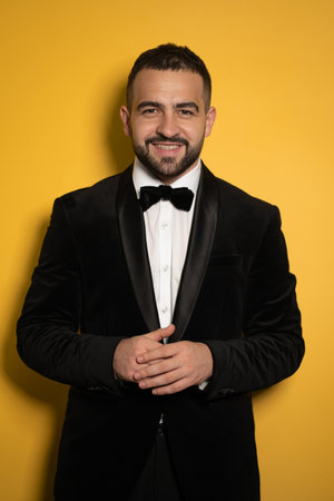 Smiling handsome man in black tuxedo with hands folded looking on camera, work profession lifestyle. Handsome young smiling caucasian man isolated on yellow background. Male fashion concept.の写真素材