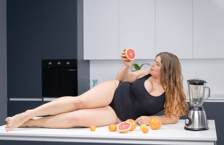 Sexy overweight girl laying on the kitchen table. An obese young sexy chubby white girl in black swimsuit at modern kitchen with fruits next to her.の写真素材