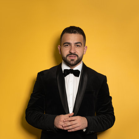 Concentrated handsome man wearing black tuxedo with hands folded looking on camera. Handsome young smiling caucasian man isolated on yellow background. Square crop.の写真素材