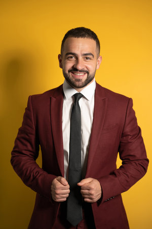 Young handsome man wearing amazing elegant burgundy suit looking on you smiling with hands holding a jacket isolated on yellow background. Men fashion concept.の写真素材