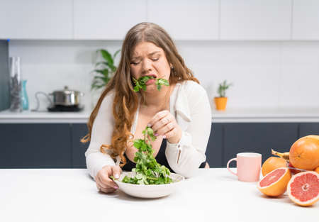 Young girl with weight problems holding fresh salad casting trying to chew it. Curvy body young woman with long blond hair sitting on modern kitchen. Dieting and nutrition concept.の写真素材