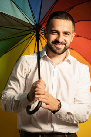 Young straight man with a rainbow umbrella behind his back. Healthy fashion lifestyle, people sincere emotions, cold season concept. Handsome guy in support of LGBTQ society.の写真素材