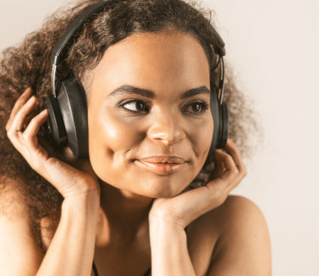 Young African-American girl listening music in headphones wearing black top isolated on grey background, emotionally move. Concept of emotions, facial expression. Square cropped.の写真素材