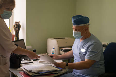 Head doctor talking to female nurse works with patient application. Male GP in protective uniform with working with documents in his office. City Hospital. May, 2020, Brovary, Ukraine.のeditorial素材