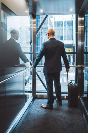 Businessman on a business trip arrived at the hotel for settlement rises in elevator standing with his back to the camera holding a suitcase by the handle. Business man looking outside of the window.の写真素材