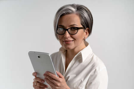 Portrait of a grey haired mature woman in white shirt and eye glasses using digital tablet. The use of technology by the elderly browsing internet shopping online on pad isolated on white background.の写真素材