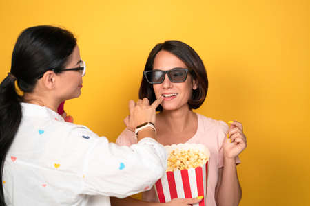 The make up artist corrects the lips of the model girl before shoot. A young woman with 3D glasses holds a box of popcorn. Two women isolated on a yellow background.の写真素材