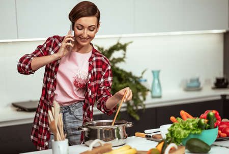 Young housewife talking on the phone while mixing a sup in a pan with a short hairstyle while...