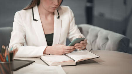 Close up beautiful office worker in white jacket making notes in her journal holding smartphone or phone in her hands sitting in front of journal. Office worker looking at her working place.の写真素材