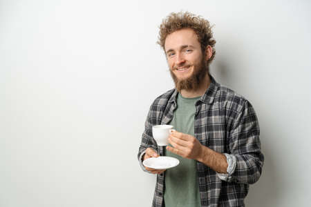 Smiling handsome man with curly hair and beard drinking coffee holding cup, wearing plaid long sleeve shirt isolated on white background.の写真素材