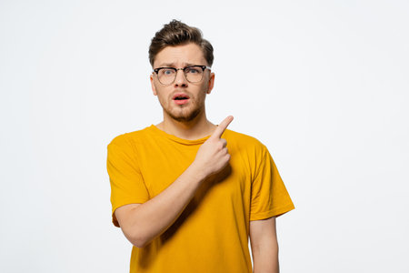 Pointing upwards to a free copy space area handsome young man in eye glasses shocked with surprise expression on his face looking at camera. Young casual man portrait isolated on white background.の写真素材