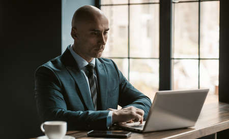 Bald middle aged businessman in business suite working on laptop next to open windows in modern...