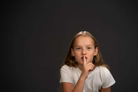 Keep quiet 8,10 years old girl holding her finger on her mouth telling everyone to keep silence wearing white t shirt smiling at the camera isolated on dark grey or black background. Copy space.の写真素材