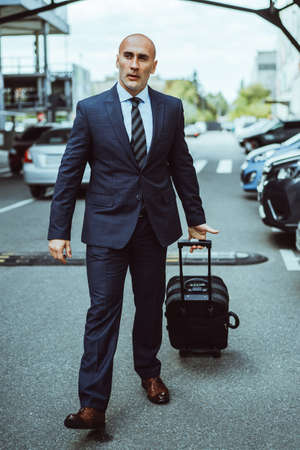 Businessman in a suit on a parking lot carrying suitcase walking thru parked cars. Businessman in suit and suitcase on business trip. Man walking outdoors with luggage determined and confident.の写真素材