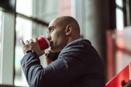Mature business man in a business suit, talking on the phone while drinking coffee using disposable paper cup standing in a office building. Business concept.の写真素材