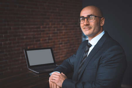 Mature bald businessman in eye glasses working alone at his desk in the office late at night, sitting half sideways with white screen on laptop and brick wall on the background.の写真素材