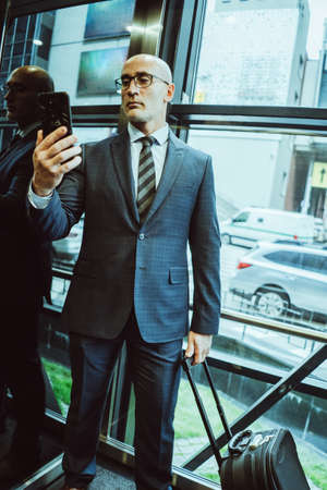 Confident businessman in suit reads information about in smartphone. Man holds mobile phone in front of him while standing next to window or elevator with busy street view in glassy wall.の写真素材
