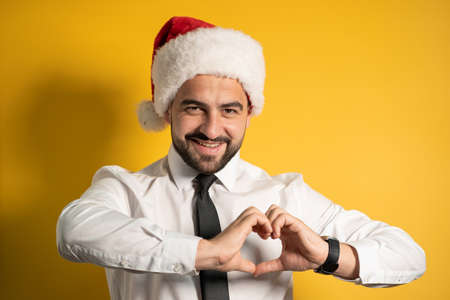 Close up. Smiling bearded man in Santa hat showing hear gesture isolated on yellow background....