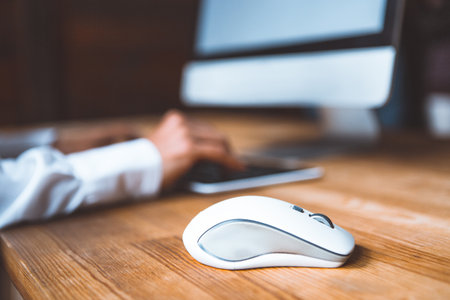 Close-up of a computer mouse on a table against the background of a working girl at a computer dressed in a white shirt. Selected focus on computer mouse on the wooden table.の写真素材