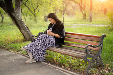 Woman of Asian ethnicity in beautiful long dress sits at park on old rusty bench and reads book using her gadget or checks social networks. Resting woman in sunny day outdoors.の写真素材