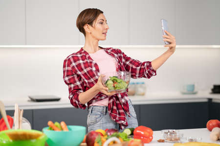 Pretty housewife taking selfie or making a video call using her smartphone while cooking fresh salad wearing a plaid shirt with a bob hair style. Healthy food leaving - vegan concept.の写真素材