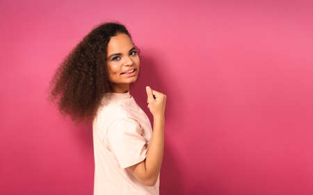 Beautiful young African American girl half turned looking positively at camera wearing peachy t-shirt showing her muscles isolated on pink background. Beauty concept.の写真素材