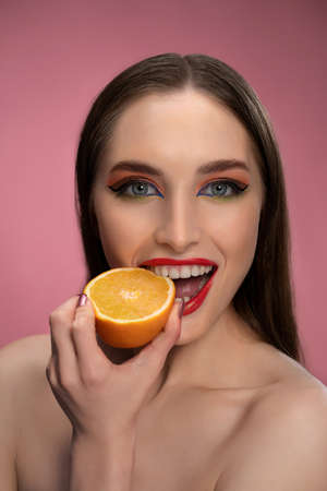Beautiful fashion girl holding orange sliced in half in her hand biting it looking at camera. Charming joyful funny lady with red lips and long hair isolated on pink background.の写真素材