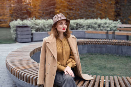 Charming young woman in beige coat and plaid hat sits on round bench waiting for her date or girlfriends. Autumn walk concept. Tinted photo.の写真素材