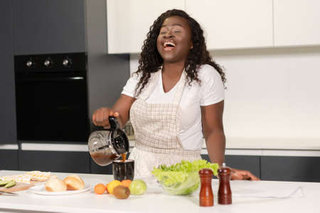 Dark Skin Woman in Apron Pours Black Coffee from a Glass Coffee Maker into a Mug. Woman Enjoy...
