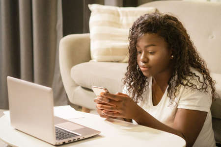 African Woman in White T-shirt is Sitting on the Floor and Texting a Message on Her Smartphone Next to Her Laptop. Close-up. Couch Background. High quality photoの写真素材