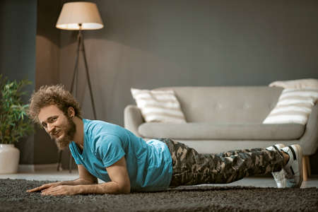 Cheerful Guy in a Blue T-shirt Does Plank Indoor. Young Man Does Up-downs to Get Fit. Medium...
