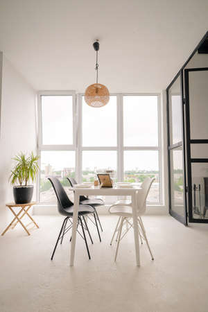White Square Dinning Table Served for 4 Persons with Black and White Chairs in the Middle of the Bright Dinning Room. White Dinning Room with Light Walls and Floor, Big Window Frame, Elements of Decor. High quality photoの写真素材