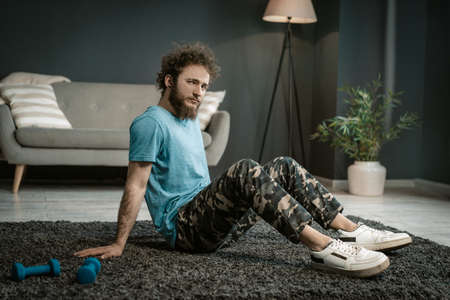 Man in a Blue T-shirt Sits on a Carpet Next to his Blue Dumbbells and Gazes at the Camera. Medium Shot. Living Room Background. High quality photoの写真素材