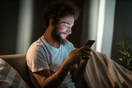 Nice Guy Texting to his Girlfriend by his Smartphone in the evening. Before Going to Bed he Sends a Mail to his Sister. Close-up. High quality photoの写真素材