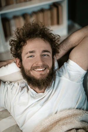 Handsome Guy with Blue Eyes, Curly Hair, a Beard and a Wide Smile Lies Under the Cover in his Bed. Top View. Close-up Portrait. Book Shelves Background. High quality photoの写真素材