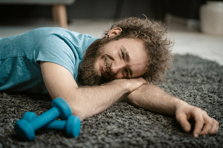Man Lies Relaxed on the Carpet Next to his Dumbbells. Curly-haired Guy is too Relaxed to Play Sports. Close-up Portrait . High quality photoの写真素材